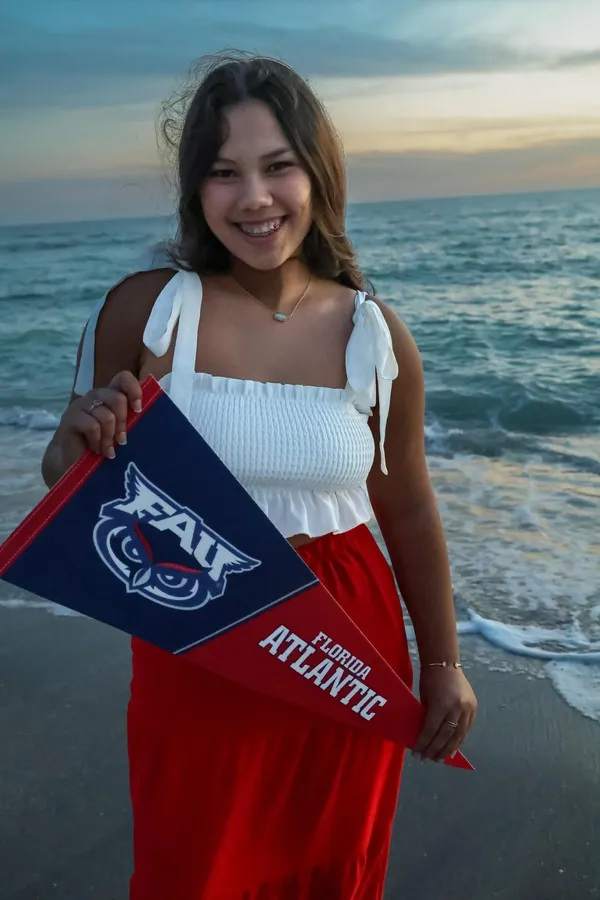 High school senior in a white top and red skirt smiling on the beach while holding a Florida Atlantic pennant at golden hour